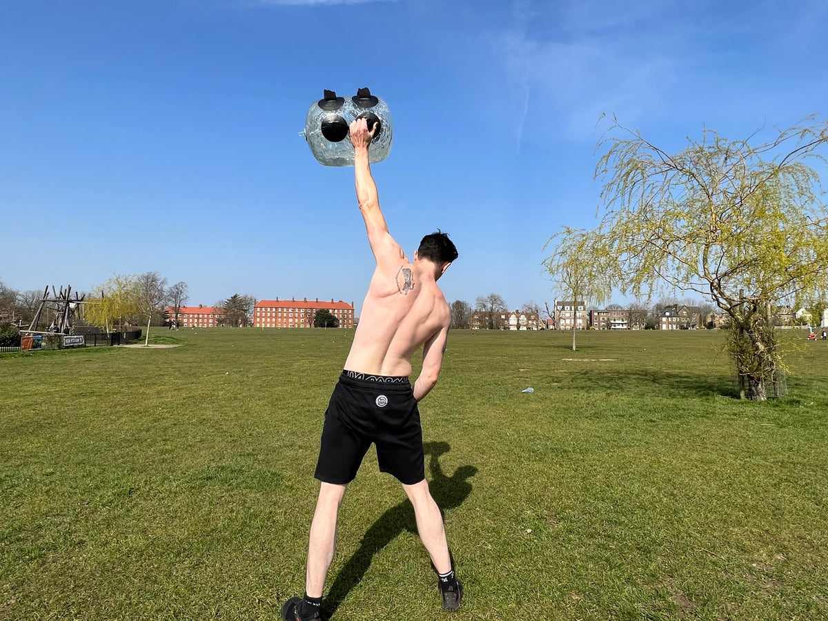 Man pressing the water-filled HELIOS weight bag overhead one-armed in a sunny park